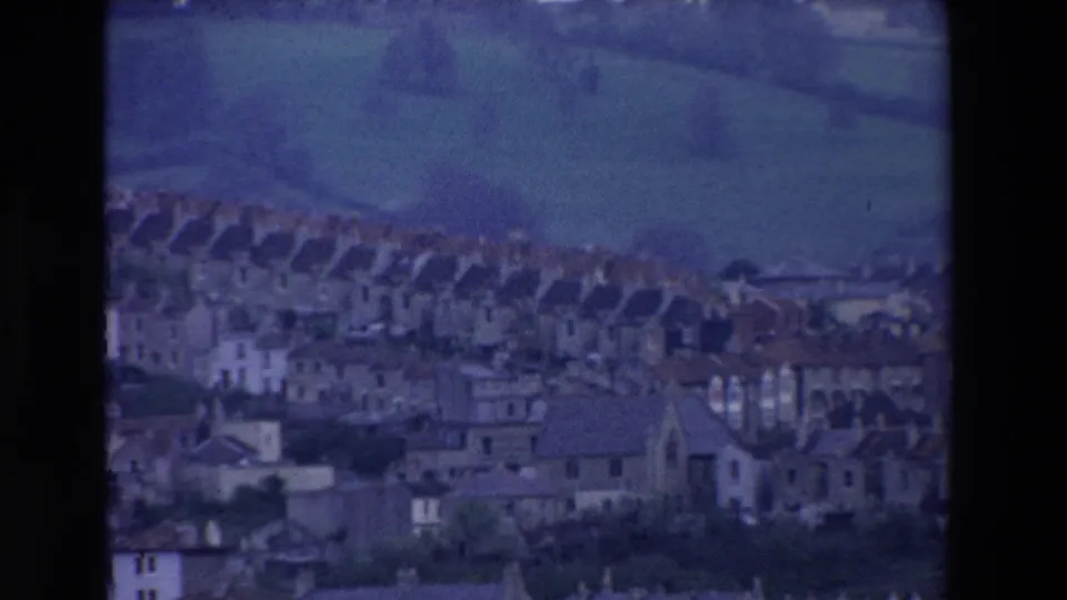 Distant 8mm view of an Irish hillside settlement in 1969, with repeated rooftops, pale building fronts, and fields rising behind the houses.