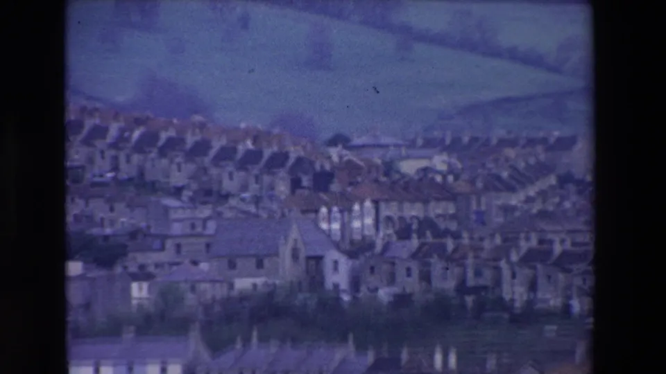 Soft 8mm frame of a hillside village in Ireland, showing dense rows of houses, a pointed roof form, green fields, and trees in the background.