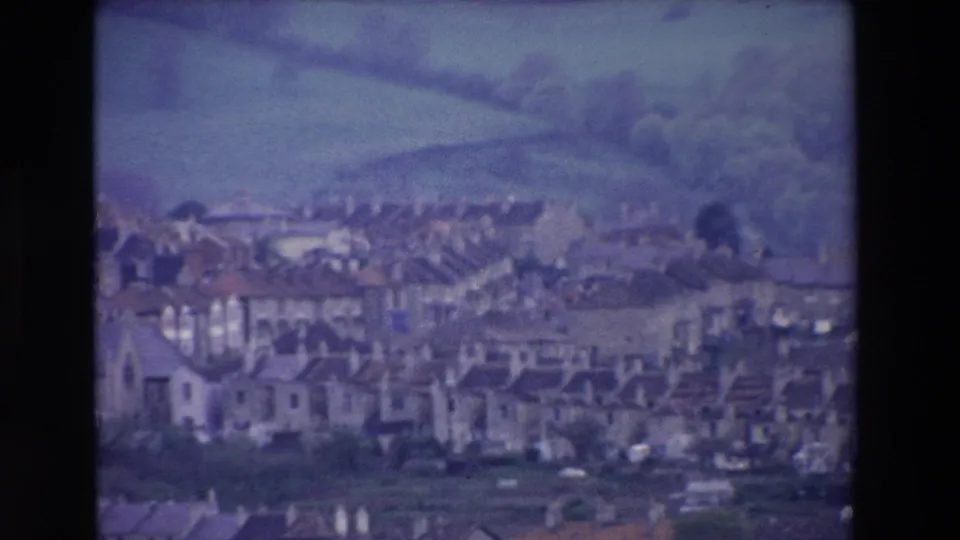 1969 Ireland home-movie frame with clustered hillside houses below rolling green fields, softened by blue-purple 8mm color shift and grain.