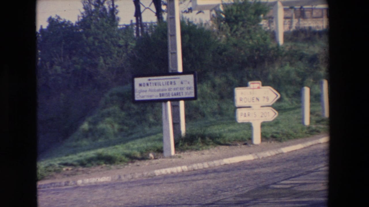 1969 8mm frame showing rural roadside signs with readable directions to Rouen and Paris beside grass, trees, and a paved road; exact location requires verification.