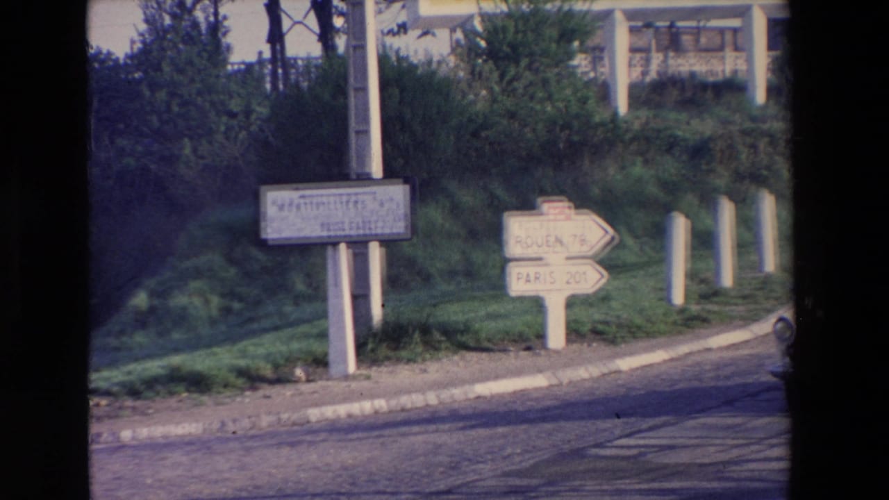 Soft 8mm frame of roadside directional signs near a paved road, including arrows toward Rouen and Paris, with shrubs and trees in the background.