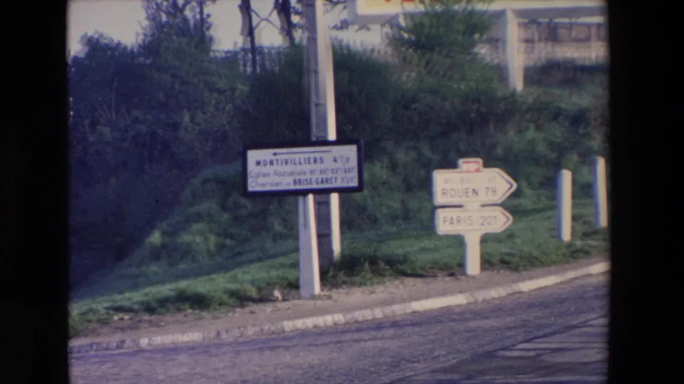 1969 8mm home-movie frame with a Montivilliers road sign and separate arrows toward Rouen and Paris beside a quiet paved road and grassy slope.