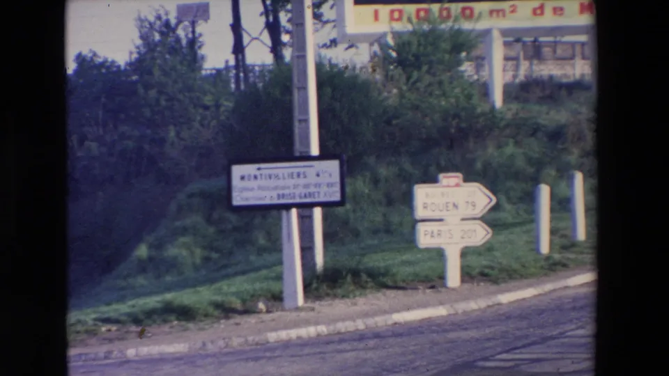 Final 8mm frame showing roadside arrows for Rouen and Paris, a Montivilliers sign, and part of an overhead sign above a grassy roadside setting.