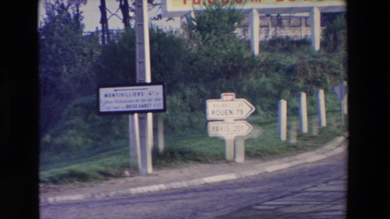 Cataloged 1969 8mm travel footage showing French directional signs for Rouen and Paris near a grassy roadside and curb.