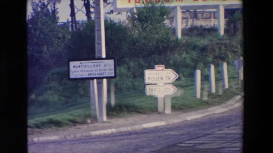 Cataloged 1969 8mm travel footage showing French directional signs for Rouen and Paris near a grassy roadside and curb.