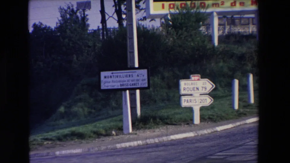 A home-movie frame holds on French road signage, roadside bollards, a curb, and dense greenery along the route.