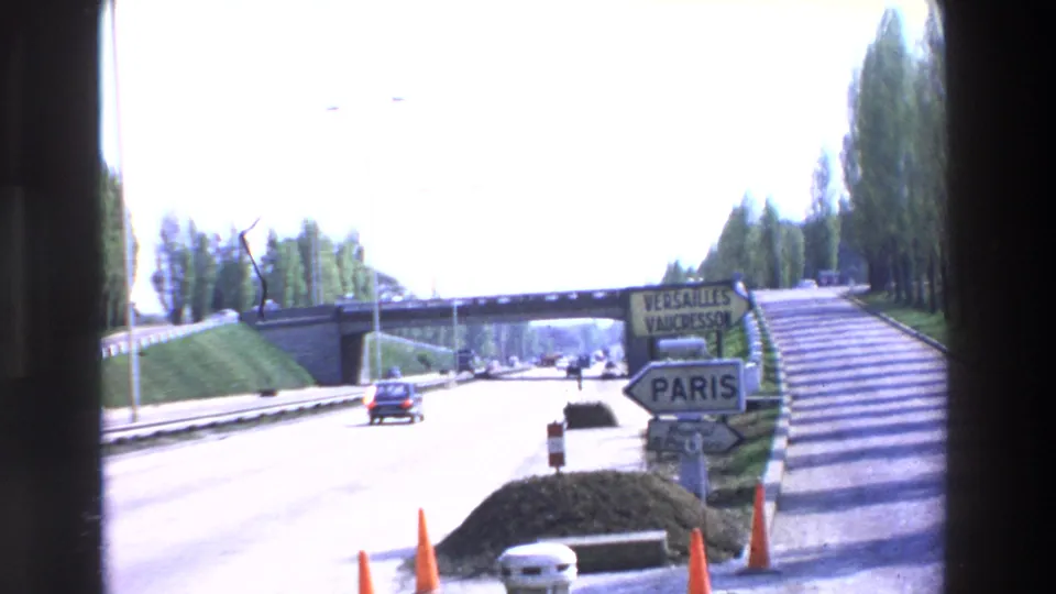 Cataloged 1969 8mm footage shows a bright highway approach with a bridge, a small car, traffic cones, and directional signs for Versailles, Vaucresson, and Paris.