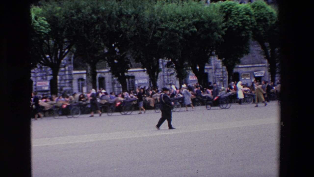 In supplied 1969 Lourdes, France footage, a man in a dark suit walks across a broad plaza as crowds gather under trees near a stone cathedral facade.