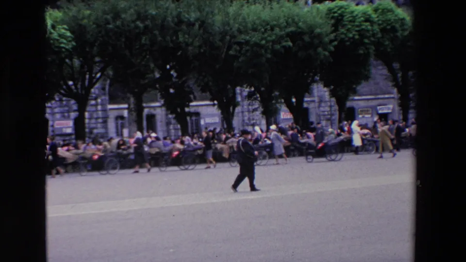In supplied 1969 Lourdes, France footage, a man in a dark suit walks across a broad plaza as crowds gather under trees near a stone cathedral facade.