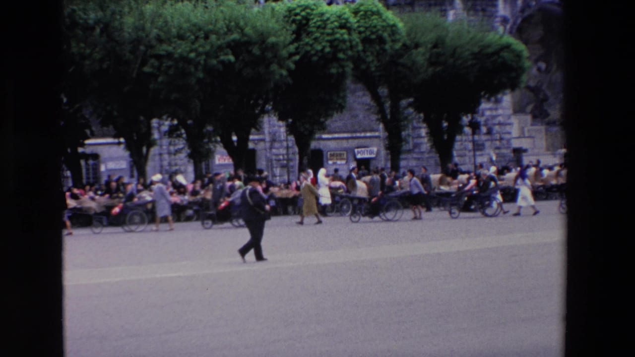 The Lourdes plaza remains busy as a suited man crosses the foreground and groups sit or move beneath trees in front of a large stone building.