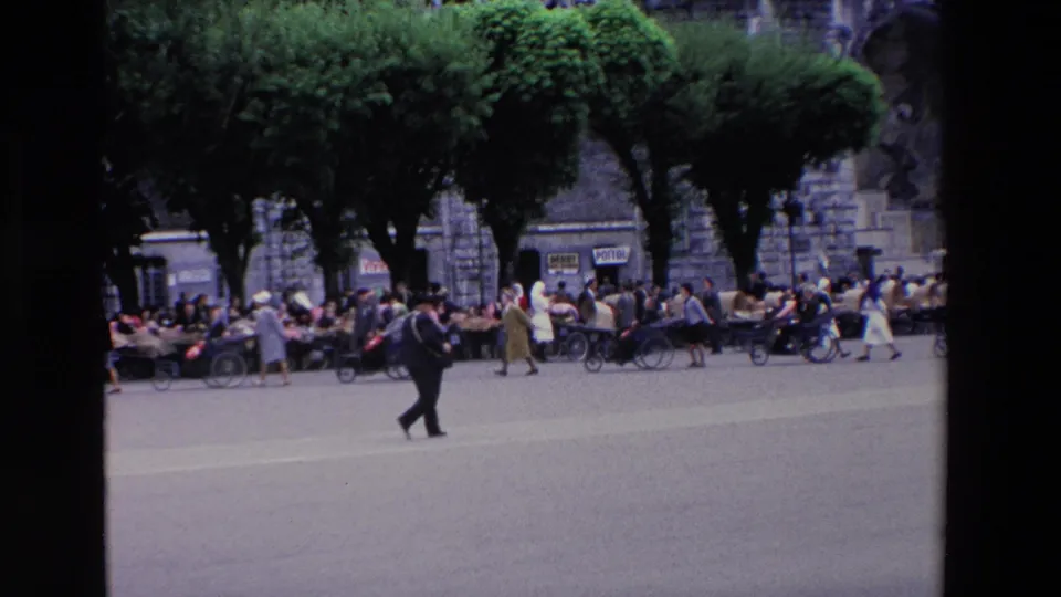 The Lourdes plaza remains busy as a suited man crosses the foreground and groups sit or move beneath trees in front of a large stone building.