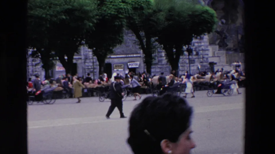 A closer handheld view shows the same crowded Lourdes square with trees, carts, pedestrians, and a foreground viewer partially entering the frame.