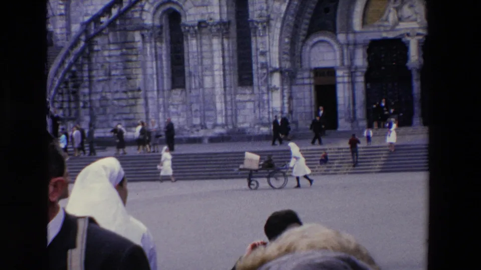 In the supplied Lourdes footage, pedestrians, white-habit figures, and hand carts move across the open area before the steps and arched entrance of a large stone cathedral.