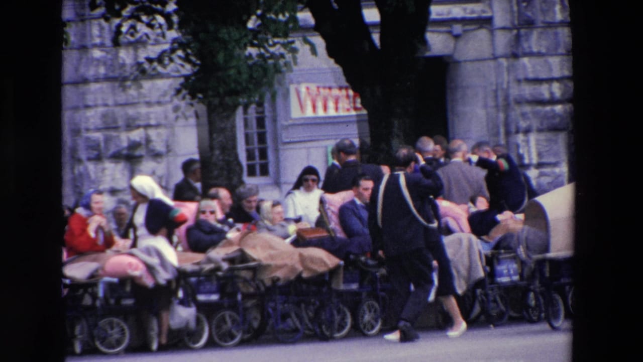 In supported 1969 Lourdes, France footage, people stand and sit near rows of vintage prams or wheelchairs outside stone buildings with partial shop signage.