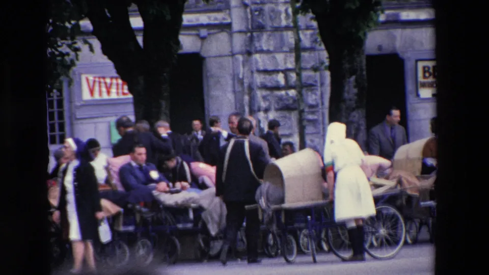 People in period clothing, including women in white veils or habits, stand among wheeled chairs and prams in the Lourdes street scene.
