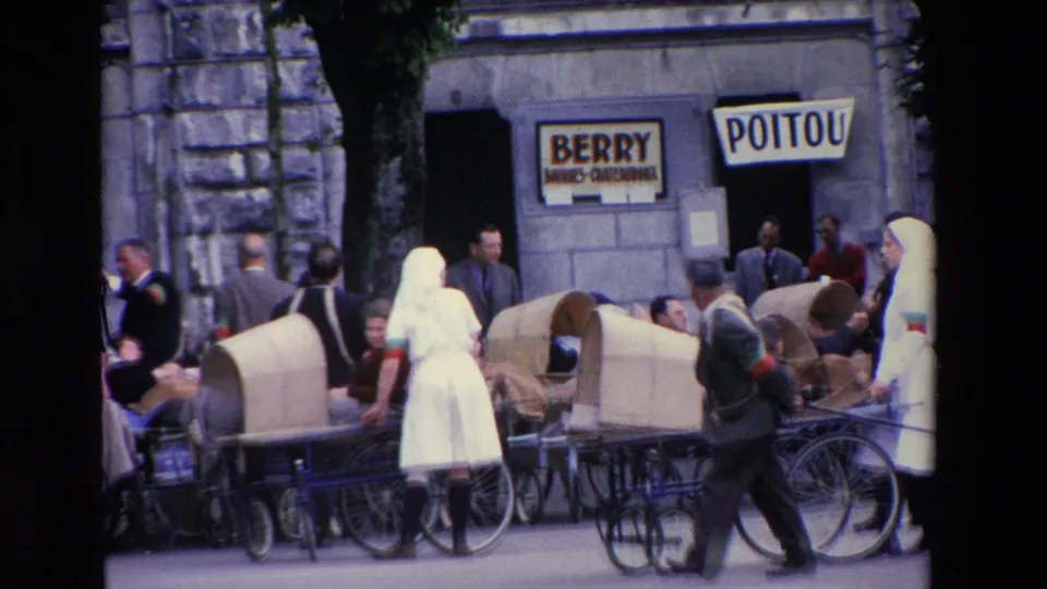 A woman in a white veil or habit stands near wheeled chairs while pedestrians move past visible BERRY and POITOU storefront signs in the 1969 Lourdes footage.