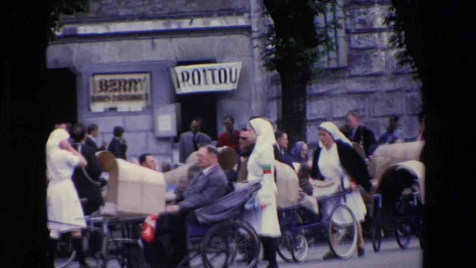 Women in white veils or habits and men in suits appear among wheeled chairs outside stone storefronts with readable BERRY and POITOU signs in the Lourdes street scene.