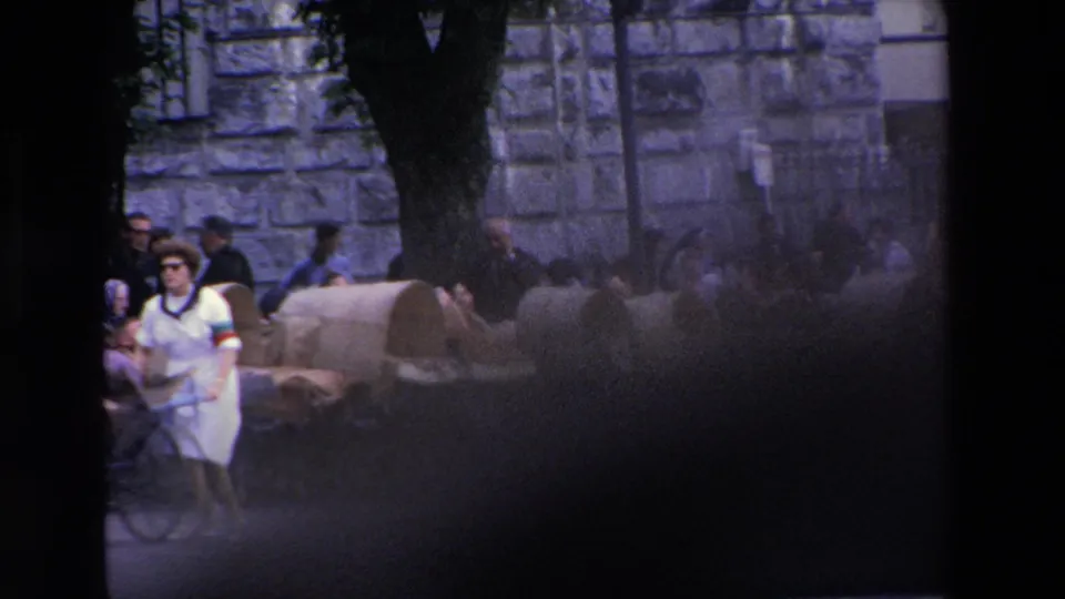 A partially obscured 8mm frame shows a nun walking beside covered carts during the Lourdes pilgrimage procession, with the stone building and street activity softened by blur and exposure variation.