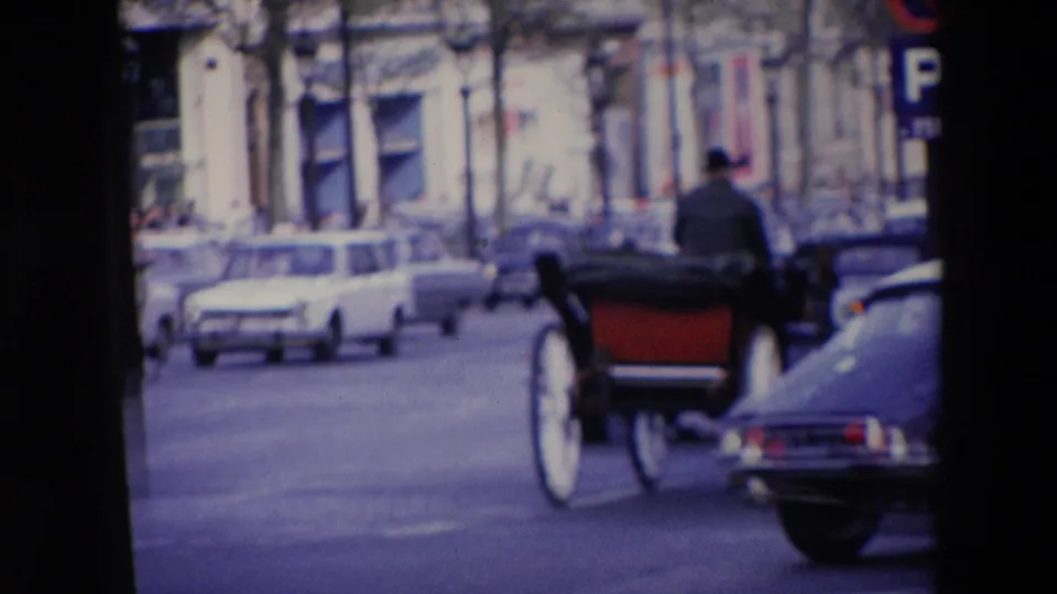 1969 home-movie view in Loudes, France, showing a horse-drawn carriage moving along a street with mid-century cars and stone-fronted buildings.