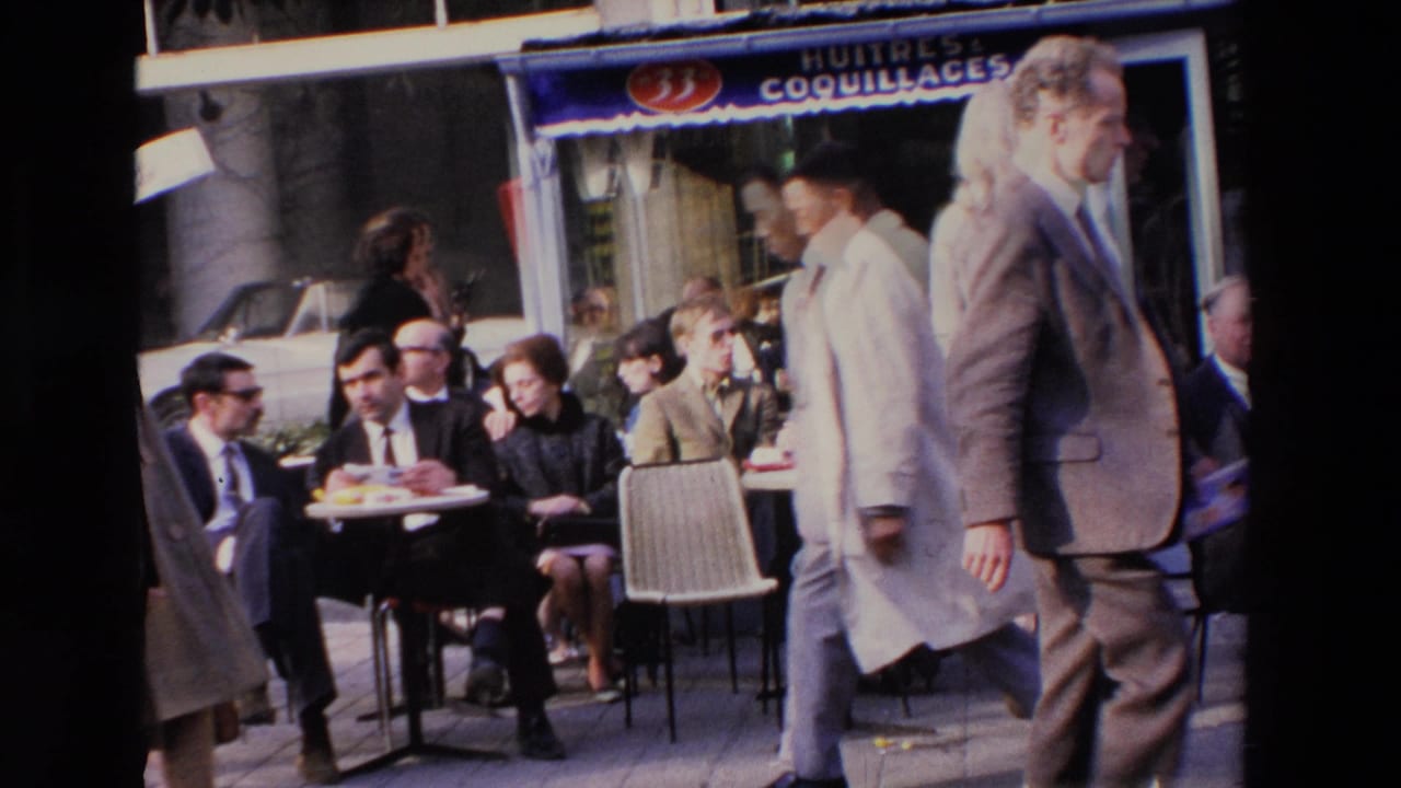Street-level 8mm frame from Loudes, France, with outdoor cafe patrons seated beneath a blue awning while pedestrians pass in front.