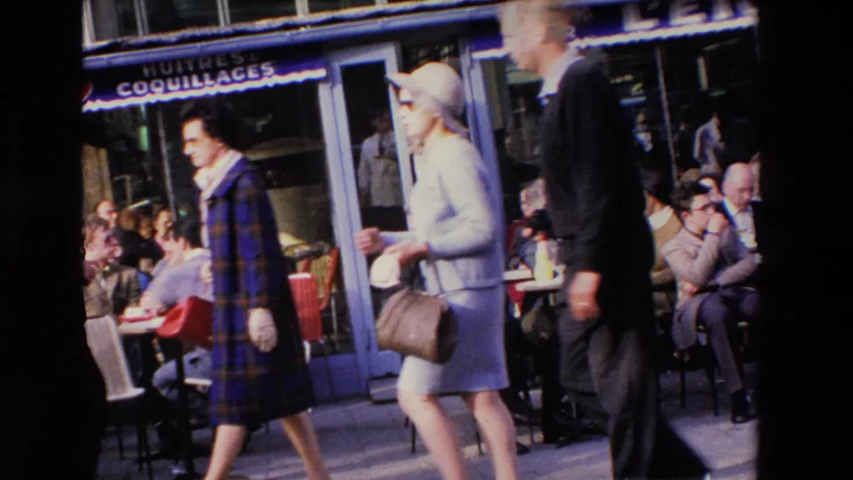 Handheld 8mm frame from Loudes, France, with pedestrians in coats and dresses walking past a cafe frontage and seated outdoor patrons.