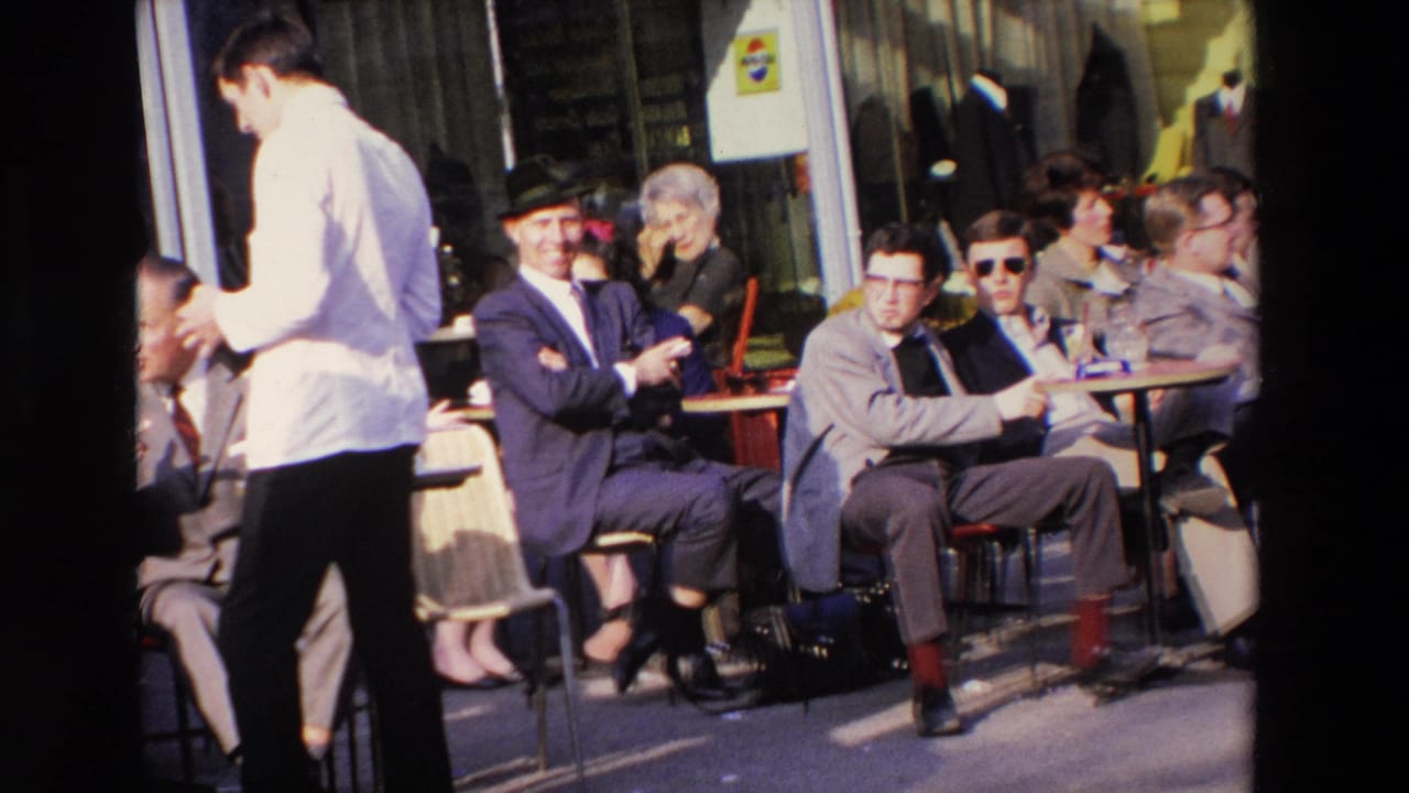 1969 French 8mm frame showing men seated at outdoor cafe tables while a waiter in a white jacket crosses the foreground on a sunlit street.