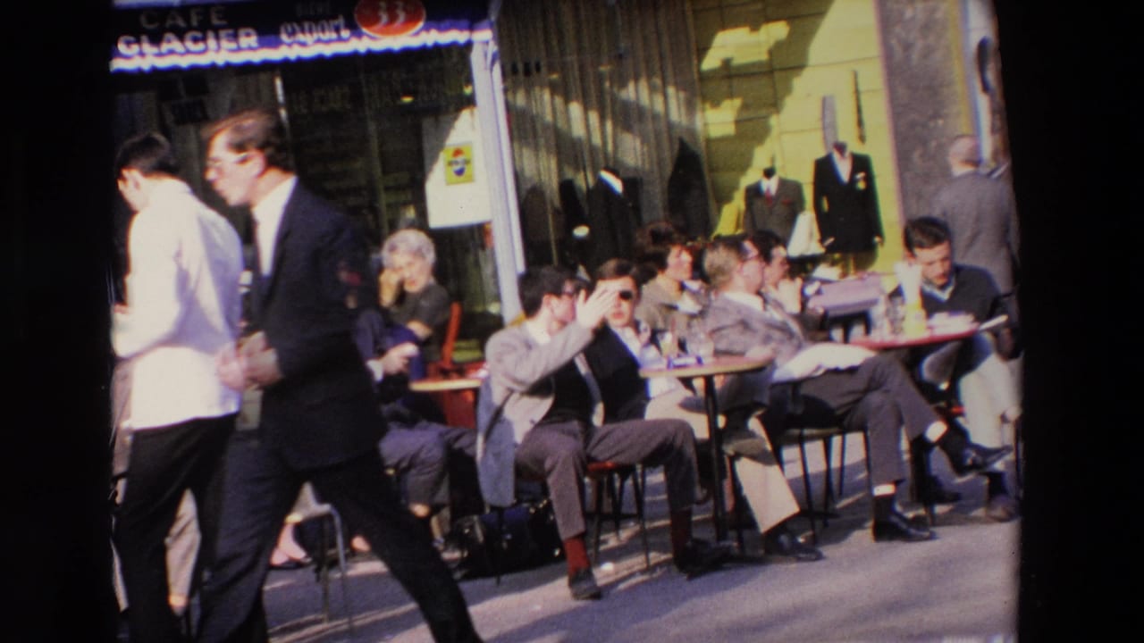 Men seated at outdoor cafe tables while pedestrians pass across the foreground.