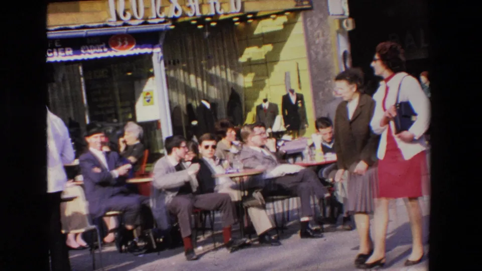 1969 French 8mm frame showing outdoor cafe patrons at tables near a storefront while two women in skirts walk through the sidewalk scene.