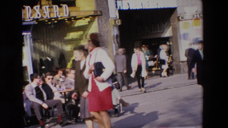 1969 French street scene with outdoor cafe seating at left, pedestrians crossing the sidewalk, storefront signage, and display windows in bright sunlight.