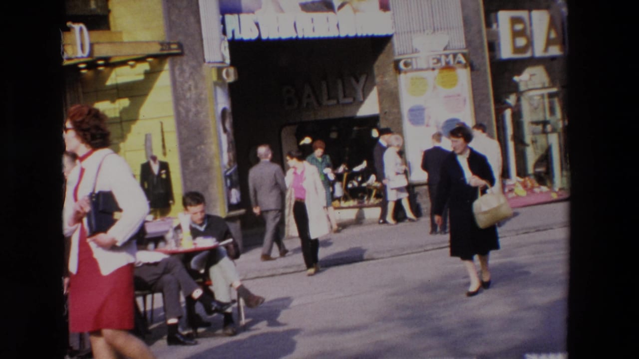 1969 home-movie frame of a busy French street with adults walking past Bally and cinema signage while a seated man occupies a sidewalk cafe table.