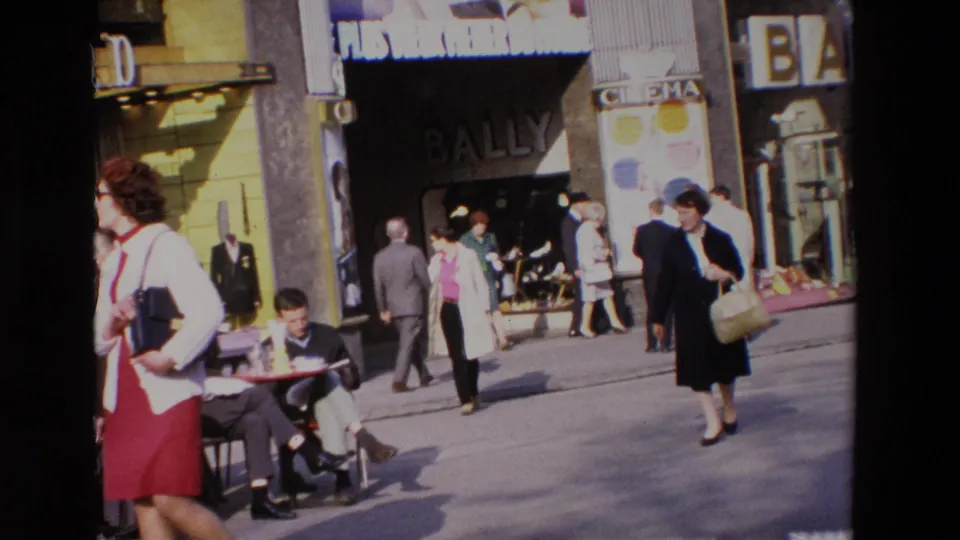 1969 home-movie frame of a busy French street with adults walking past Bally and cinema signage while a seated man occupies a sidewalk cafe table.