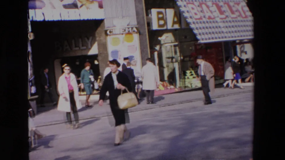 1969 home-movie frame showing women in period coats crossing a commercial street with Bally lettering, cinema signage, and pedestrians in the background.