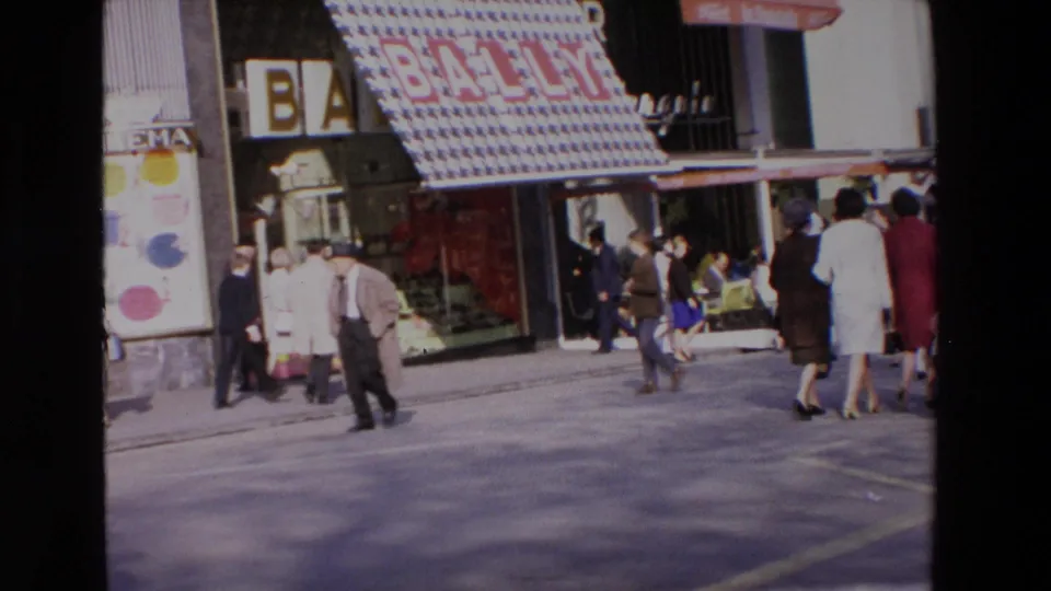 1969 home-movie frame of adults walking and pausing outside a Bally storefront, with colorful signage, shop windows, and a commercial sidewalk visible.