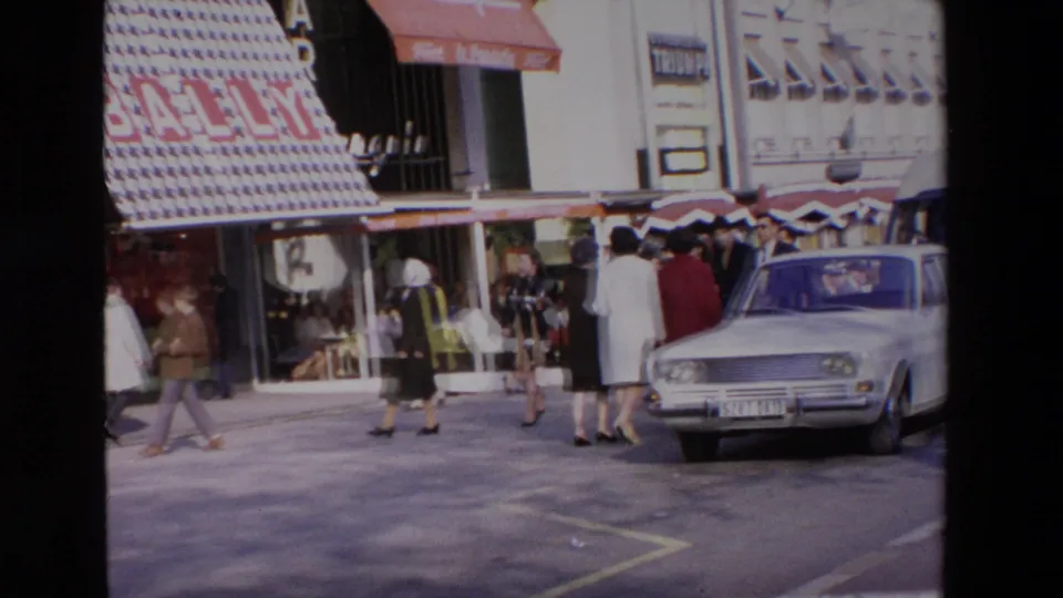 1969 home-movie frame showing a compact mid-century car at the curb or roadway beside pedestrians, storefront awnings, and the Bally shop frontage in the stated French location.