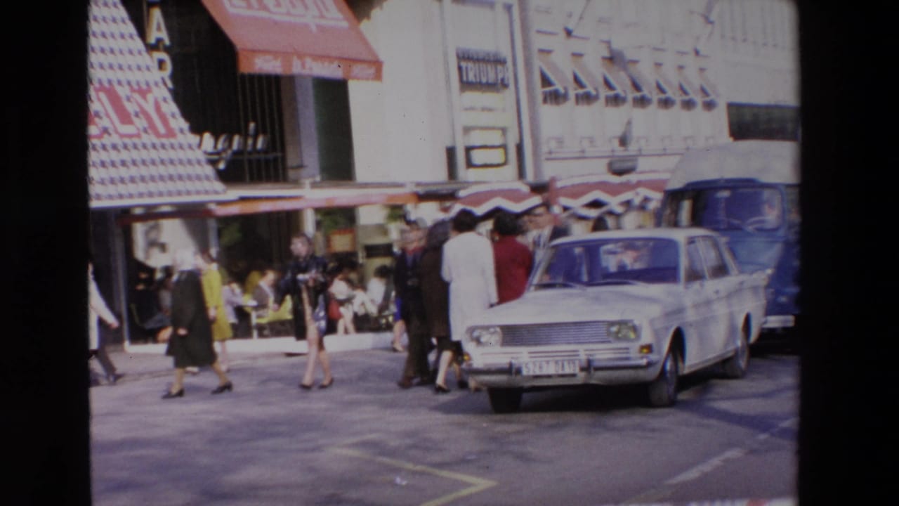 In metadata-identified Loudes, France, a late-1960s street scene shows pedestrians in period clothing near a white sedan, shopfront awnings, and storefront signage.