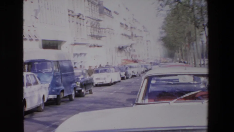 The frame shows a sunlit late-1960s street with a blue van, several parked cars, shopfronts, and pedestrians receding along the sidewalk.