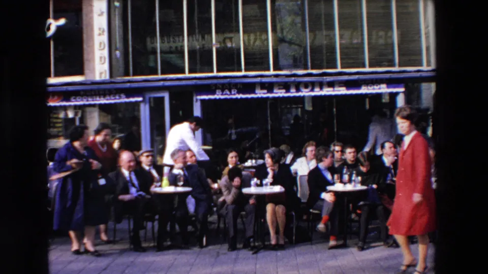 A crowded outdoor cafe in the metadata-identified French town shows seated patrons, a waiter in white, storefront signage, and a woman in a red coat walking past.