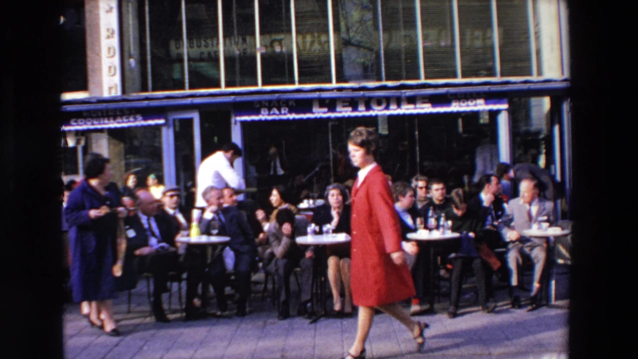 1969 home-movie frame from Loudes, France showing an outdoor cafe with seated adults, a waiter in a white jacket, and a woman in red passing in front of L'ETOILE signage.