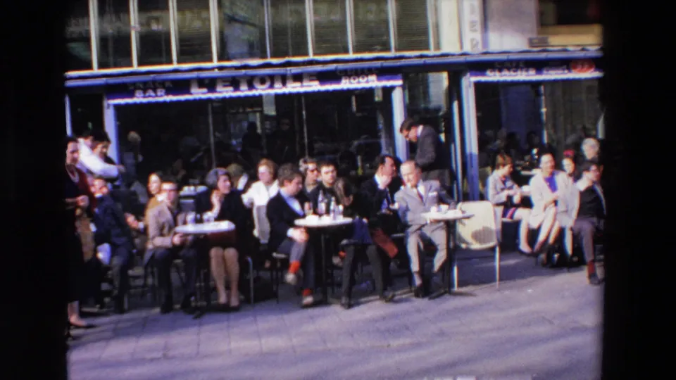 Home-movie frame of a crowded sidewalk cafe in France, with adults in period clothing seated at small tables and a waiter working near the storefront.