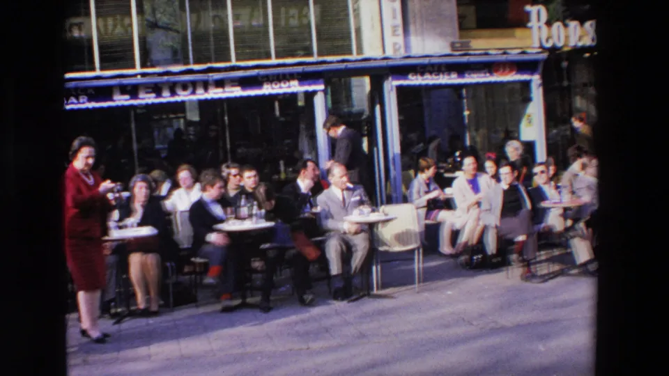 Late-1960s cafe frontage in Loudes, France with seated patrons, a woman in red near the left side, visible snack bar signage, and pedestrians along the sidewalk.