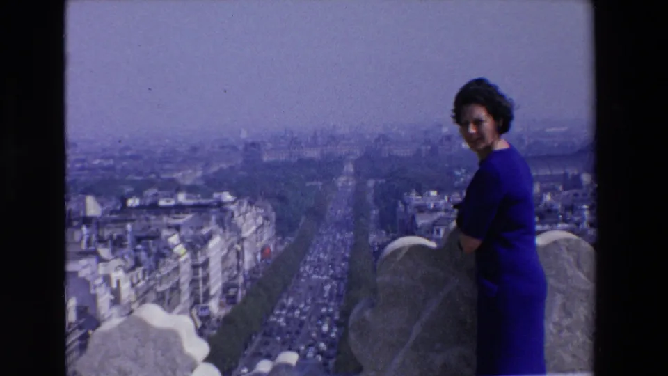 1969 France home-movie frame of a woman in a blue dress glancing toward the camera from a stone overlook above a busy urban boulevard.