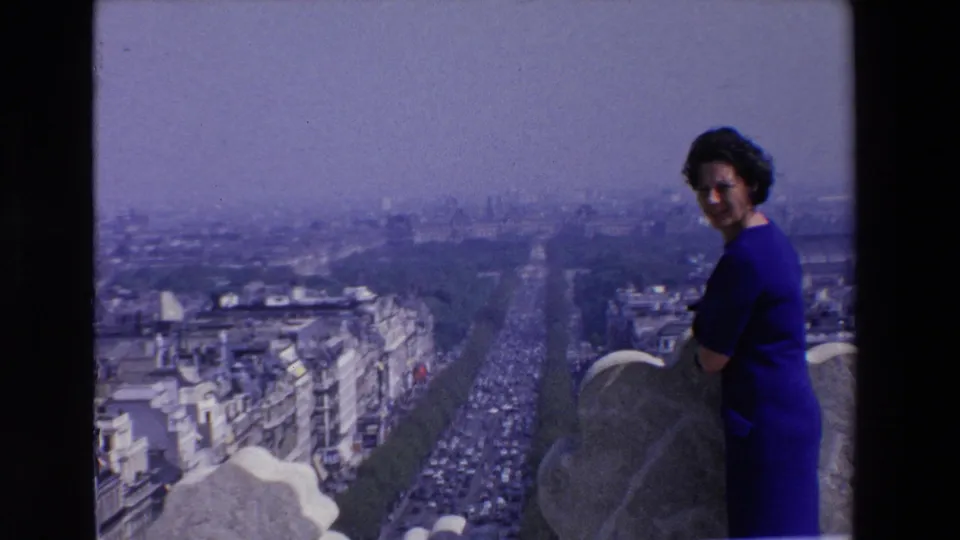 Soft 8mm view of a woman in blue at a high French city viewpoint, with traffic, rooftops, and a hazy skyline stretching behind her.