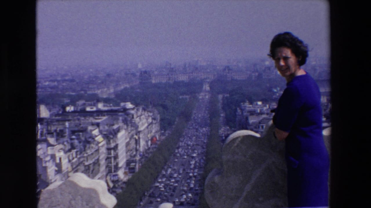 Adult woman in a blue dress stands beside a rooftop parapet overlooking a broad French city boulevard in 1969 home-movie footage.