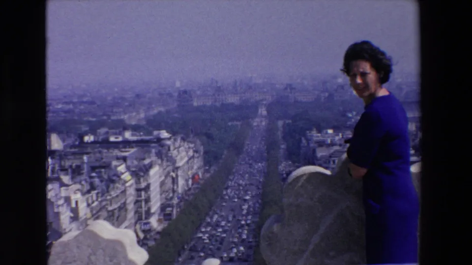 Adult woman in a blue dress stands beside a rooftop parapet overlooking a broad French city boulevard in 1969 home-movie footage.