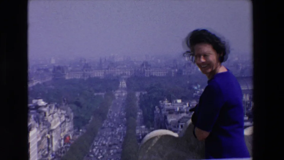 The woman turns and smiles toward the camera from a high rooftop, with a hazy French cityscape and traffic-filled boulevard behind her.