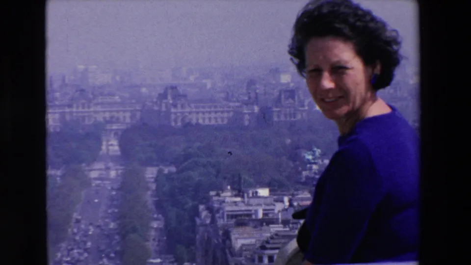 Close rooftop view of the woman in a blue dress looking toward the camera, with layered city buildings, trees, and a wide boulevard in the background.