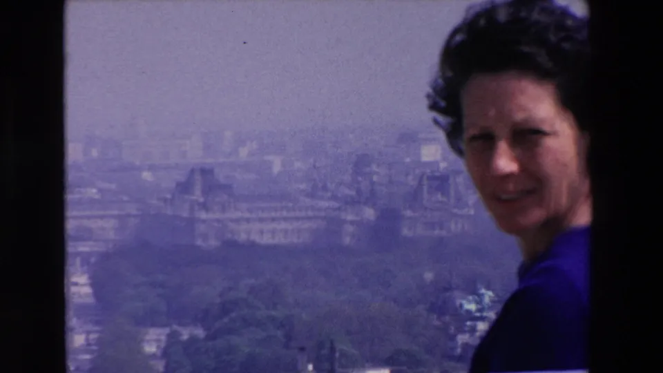 The frame moves closer to the woman's face and blue dress at the rooftop edge, with soft-focus urban buildings and greenery behind her.