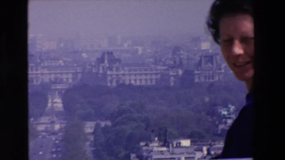 Adult woman in a dark purple top smiles near the camera at an elevated French city viewpoint, with hazy buildings and trees in the background.