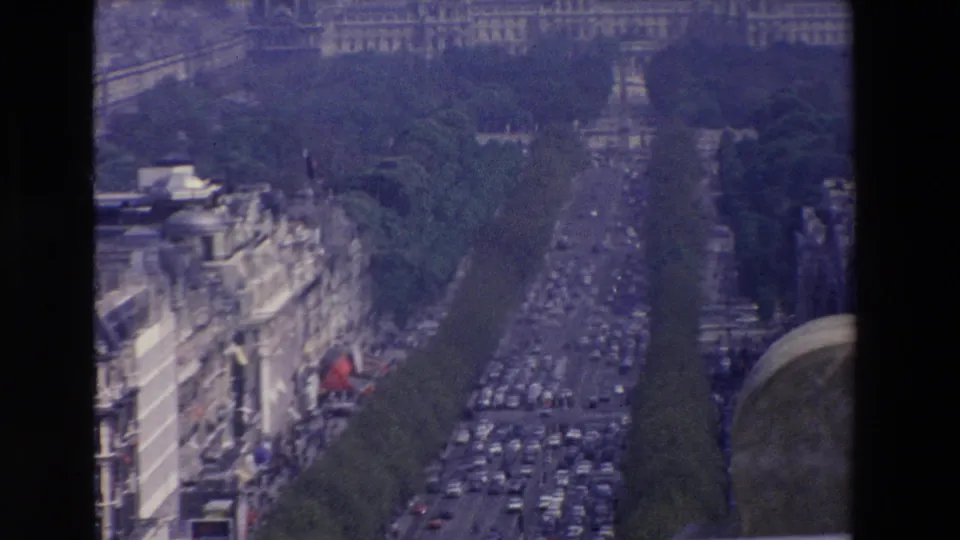 Elevated home-movie frame looking down a wide French boulevard with dense car traffic, rows of trees, ornate buildings, and a stone foreground edge.