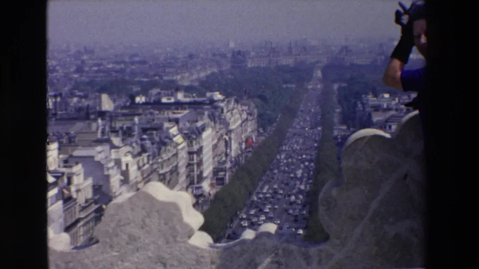 Wide elevated view over a busy French boulevard and surrounding city buildings, with stonework in the foreground and a partially visible adult at the right edge.